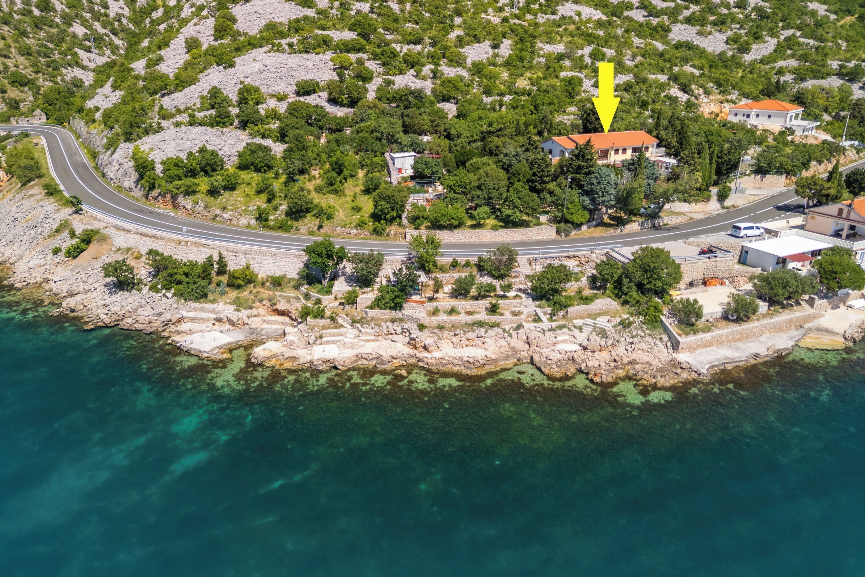 Vue aérienne d’une zone côtière avec une route sinueuse, des eaux turquoise et limpides, une côte rocheuse et une flèche jaune pointant vers une maison avec un toit rouge.