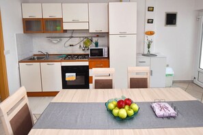 A modern kitchen with white cabinets, a gas stove, an oven, and a microwave. There is a dining table with chairs and a bowl of fruit. - Apartment Kety (Novalja)