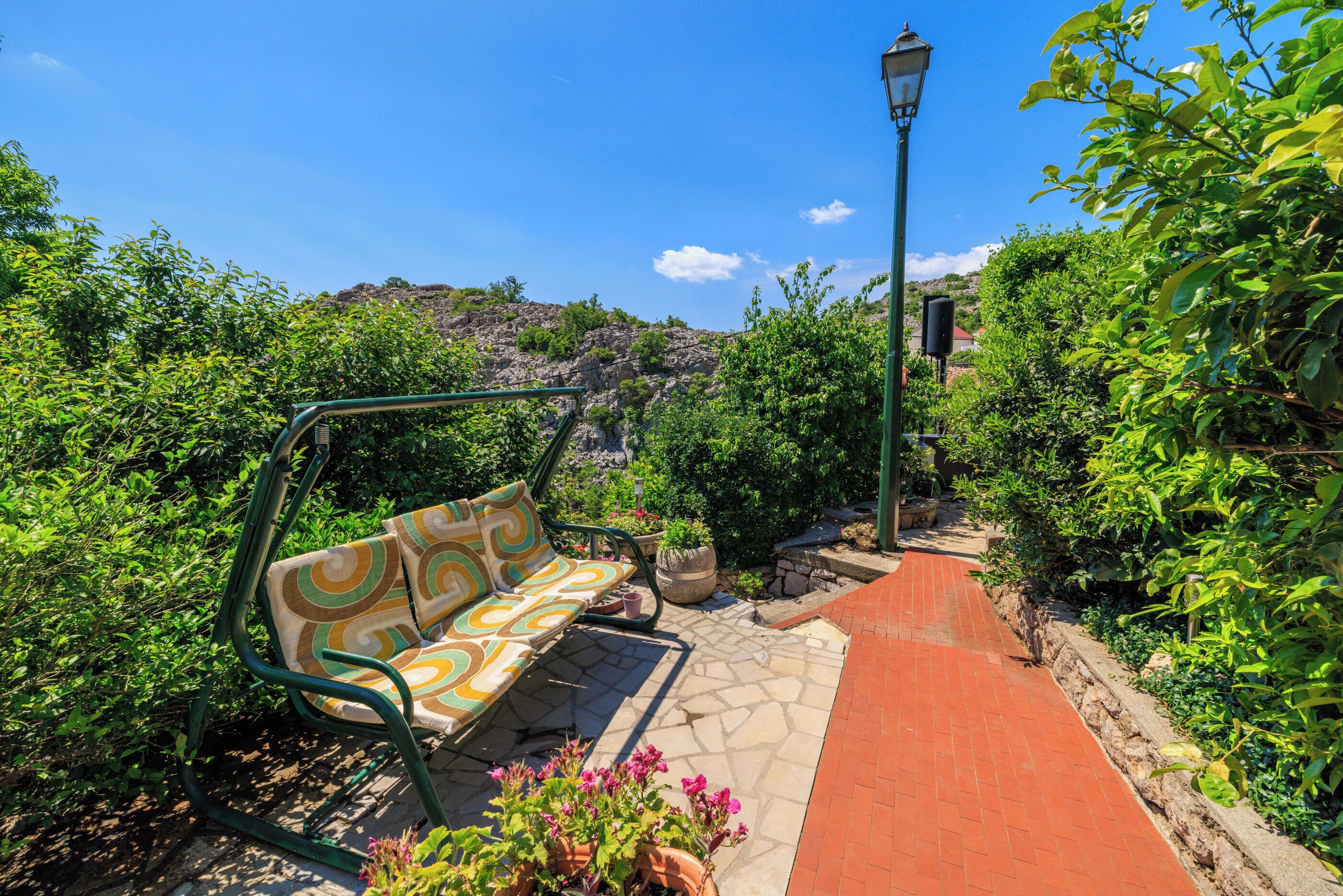 A bench with a patterned cushion, a paved walkway, and a lamp post in a garden setting.