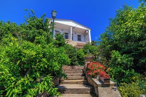 Ein Gebäude mit Balkon, umgeben von üppiger Vegetation und einer Steintreppe, die darauf führt.