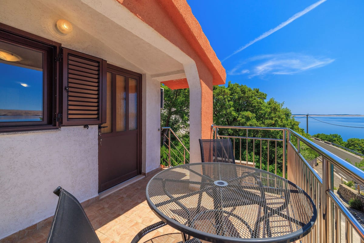 A balcony with a round metal table and chairs, a door with shutters, and a view of the sea.