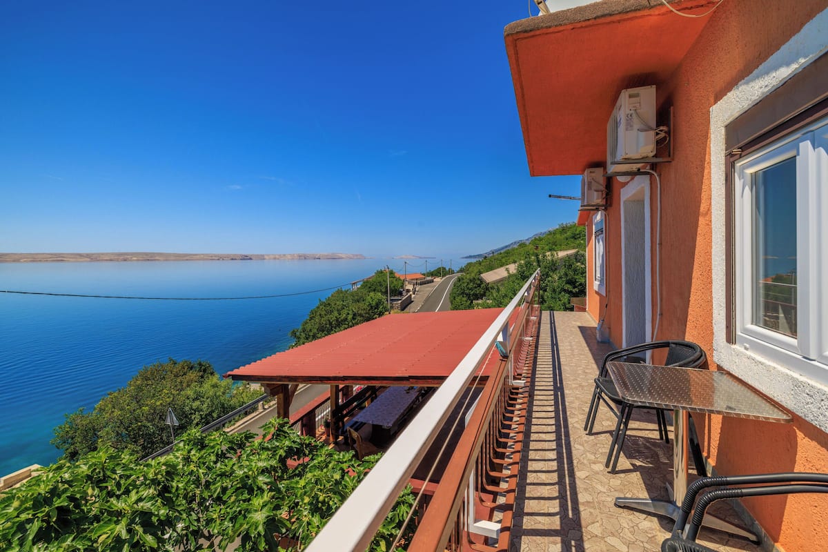 A balcony with a red roof overlooking a body of water, a table, and two chairs.