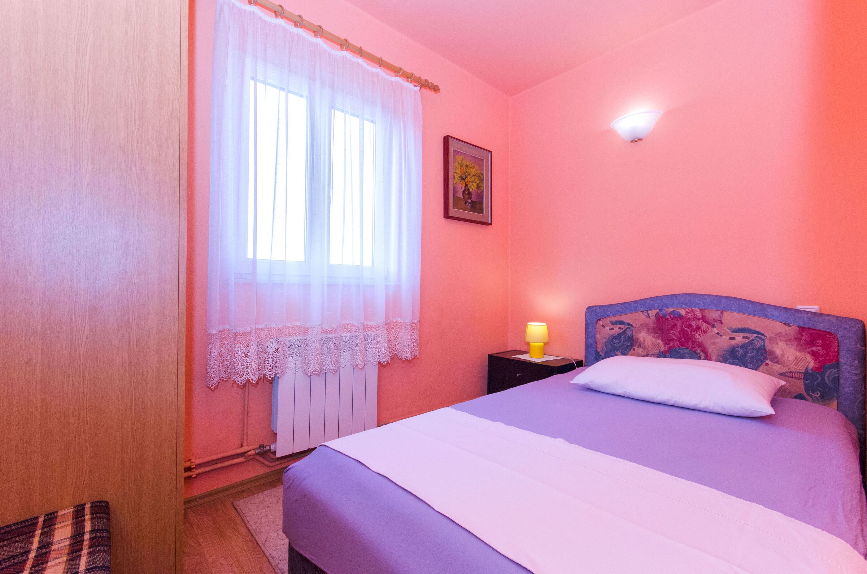 A bedroom with a bed, a bedside table with a lamp, a window with sheer curtains, and a framed picture on the wall.