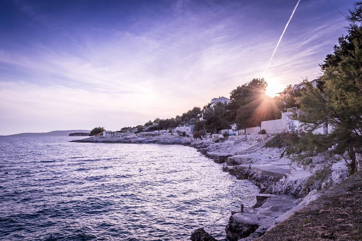 Paesaggio costiero con una costa rocciosa, cielo sereno e il sole che tramonta o sorge all'orizzonte.