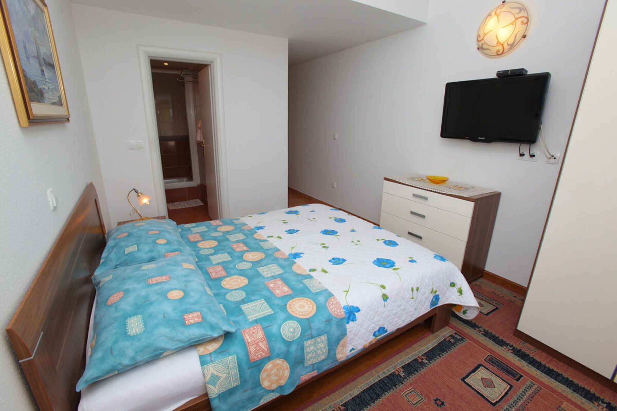 Ein Schlafzimmer mit einem Bett, einem Nachttisch, einem an der Wand befestigten Fernseher und einer Tür zu einem anderen Raum.