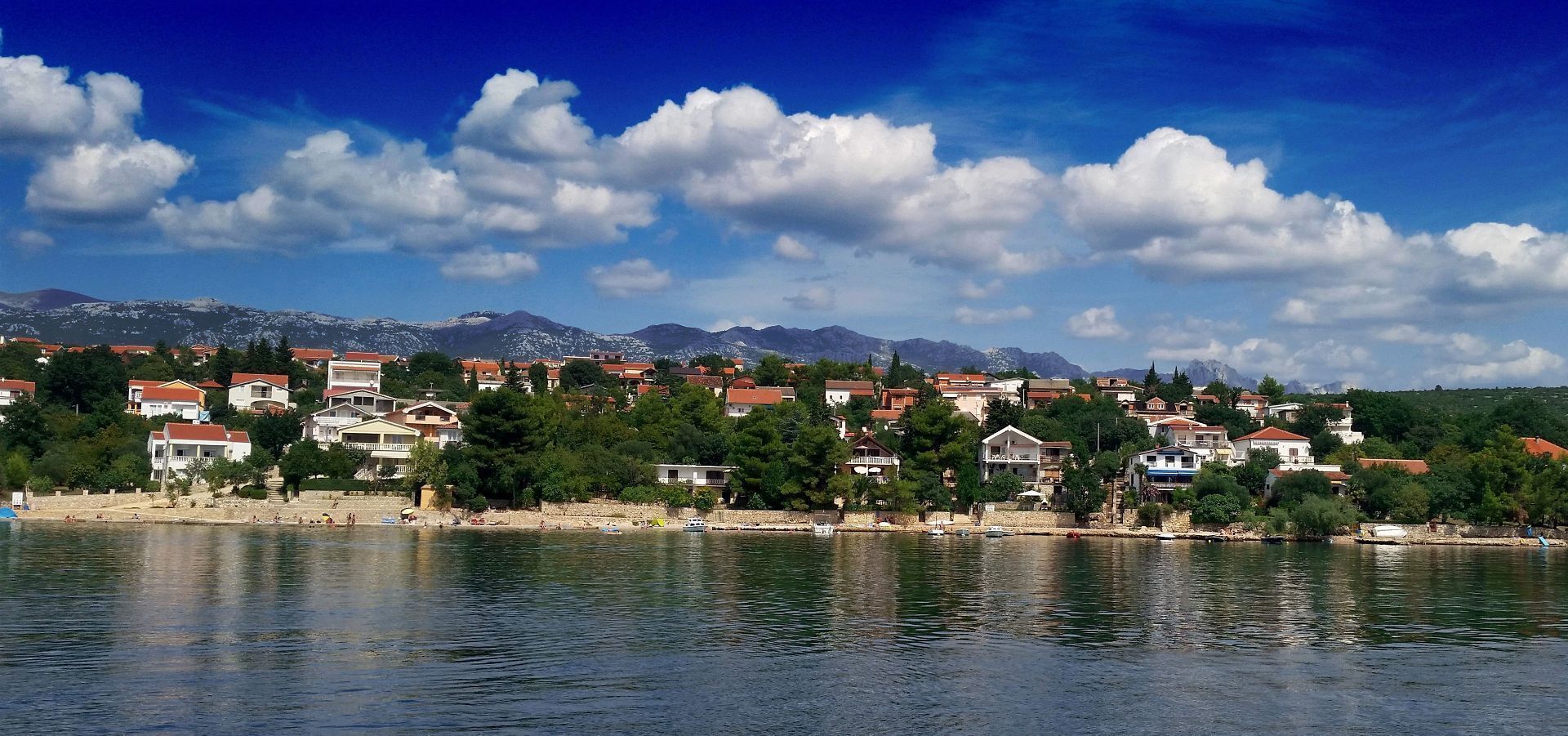 A coastal town with buildings along the shoreline, a clear sky, and mountains in the background.