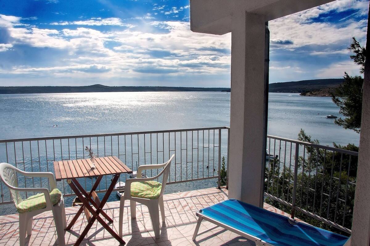 A balcony with a view of a lake, a table, two chairs, and a sun lounger.