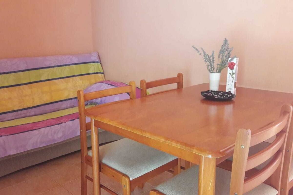 A room with a wooden dining table, a colorful sofa, and a small table with a vase of flowers.