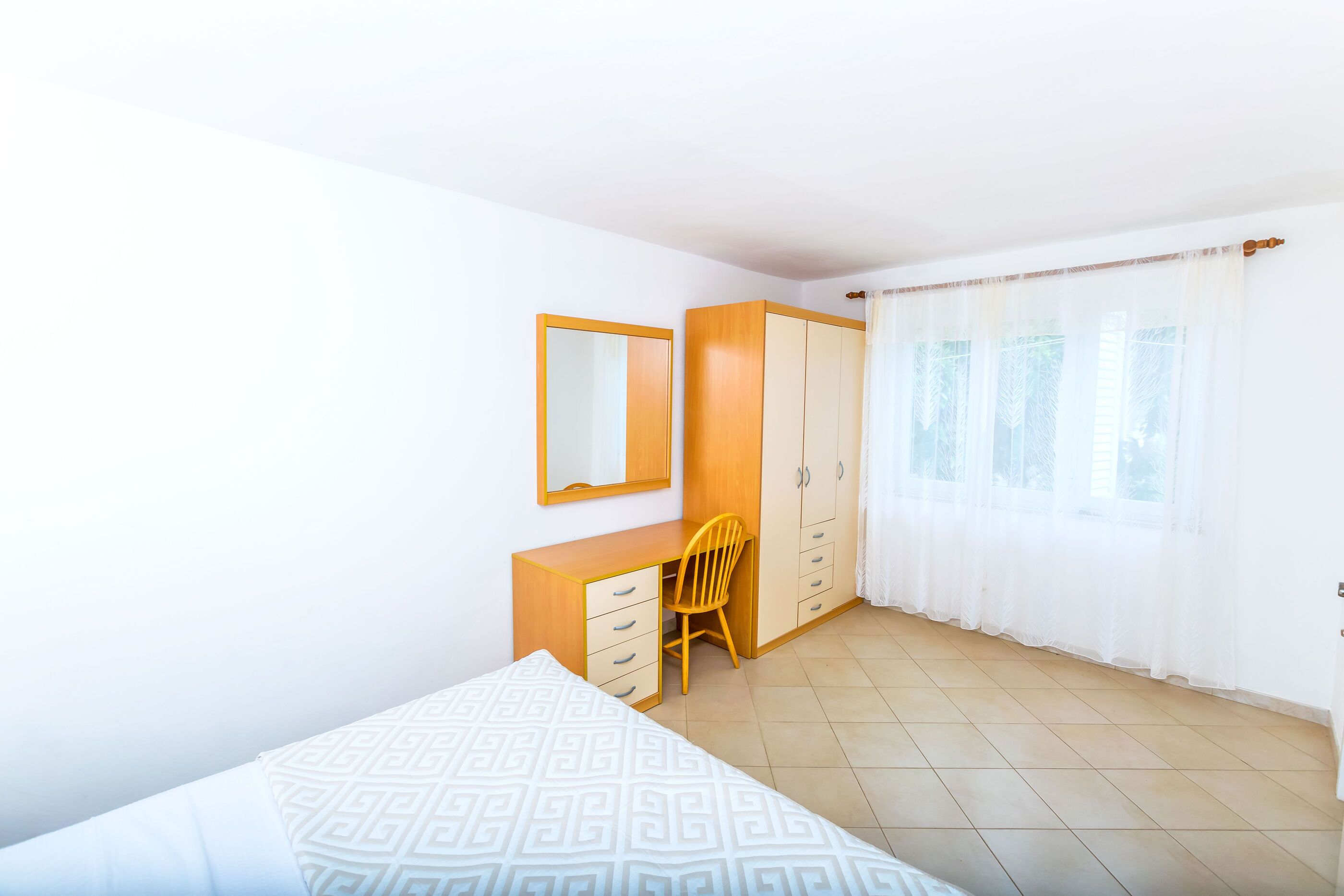 A bedroom with a bed, a wooden desk, a chair, a wardrobe, and a window with curtains.