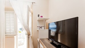 A modern living room with a flat-screen TV on a wooden console, a white shelf with decorative items, and a view of a balcony with white blinds. - Apartments Dado (Lumbarda)