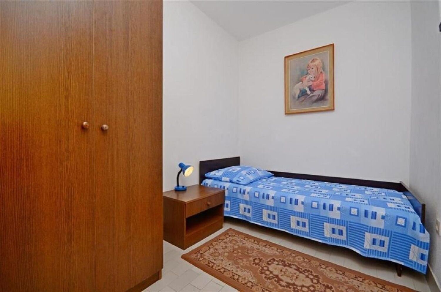 Ein Schlafzimmer mit einem Holzschrank, einem Bett mit blumigem Überwurf, einem Nachttisch mit Lampe, einem gerahmten Bild an der Wand und einem gemusterten Teppich.