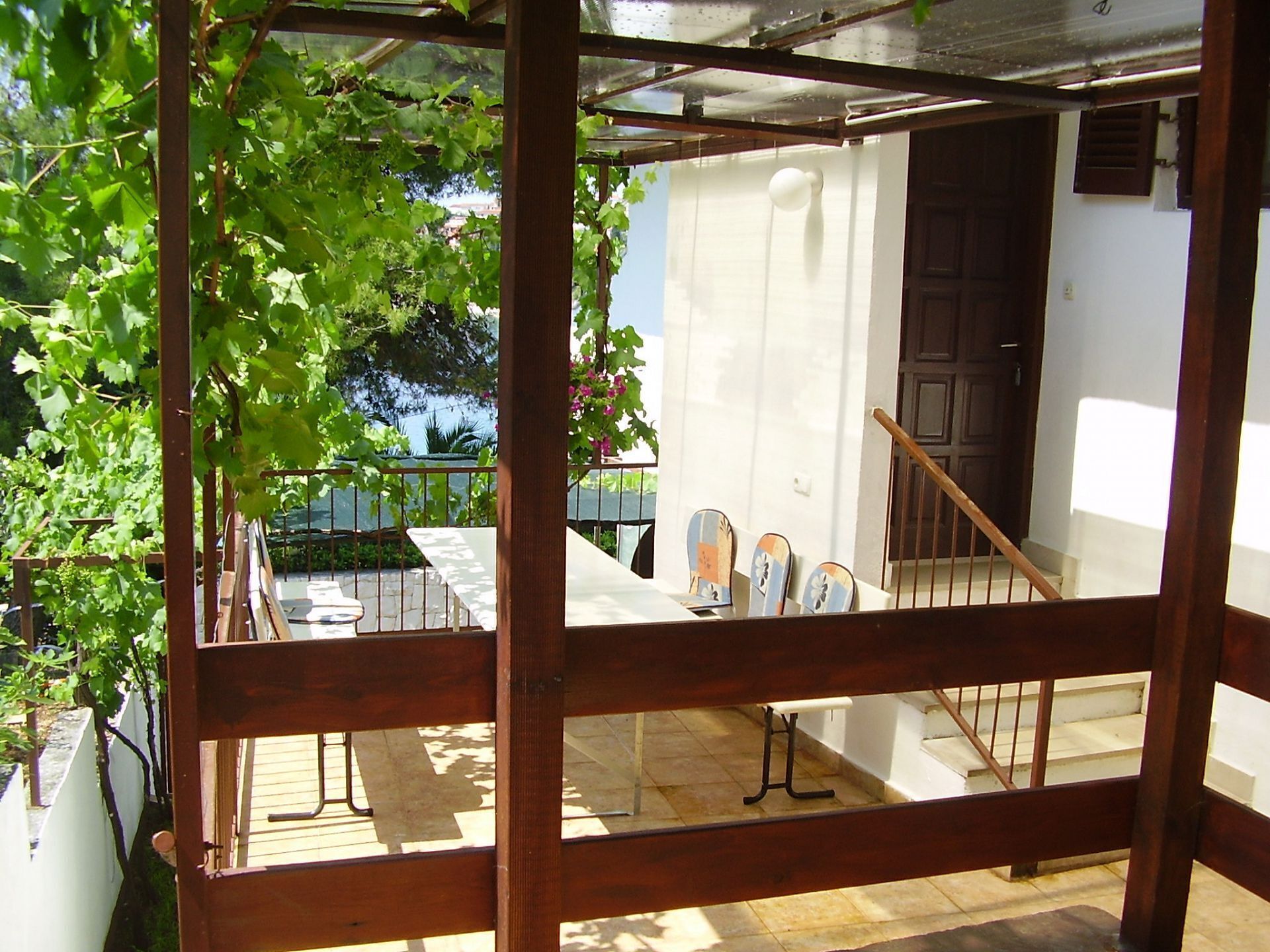 Een overdekt buitengebied met een houten pergola, een trap en uitzicht op een wit gebouw.