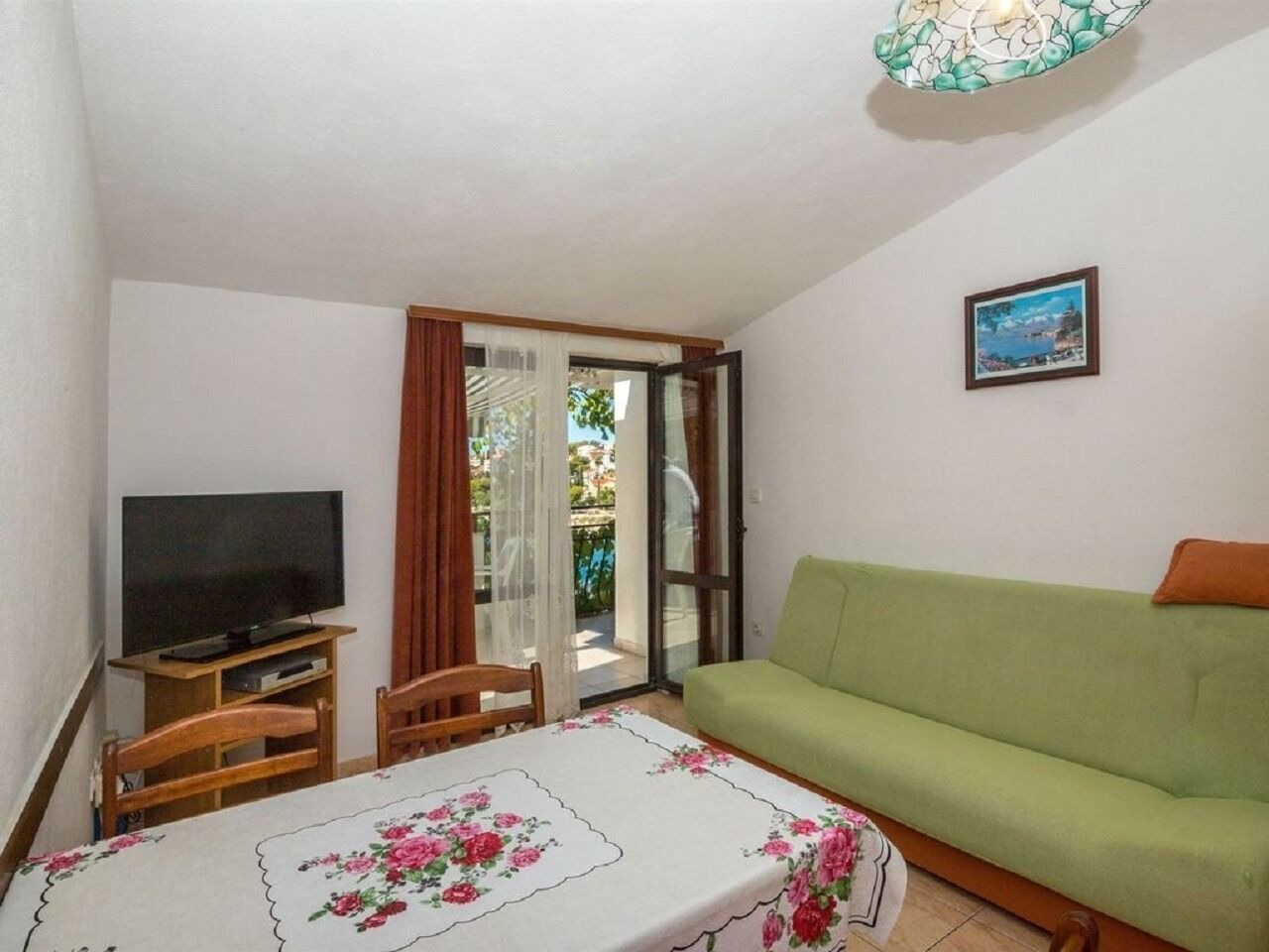 A living room with a green sofa, a television on a wooden stand, a table with a floral tablecloth, and a sliding glass door leading to an outdoor area.