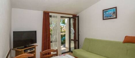 A living room with a green sofa, a television on a wooden stand, a table with a floral tablecloth, and a sliding glass door leading to an outdoor area.
