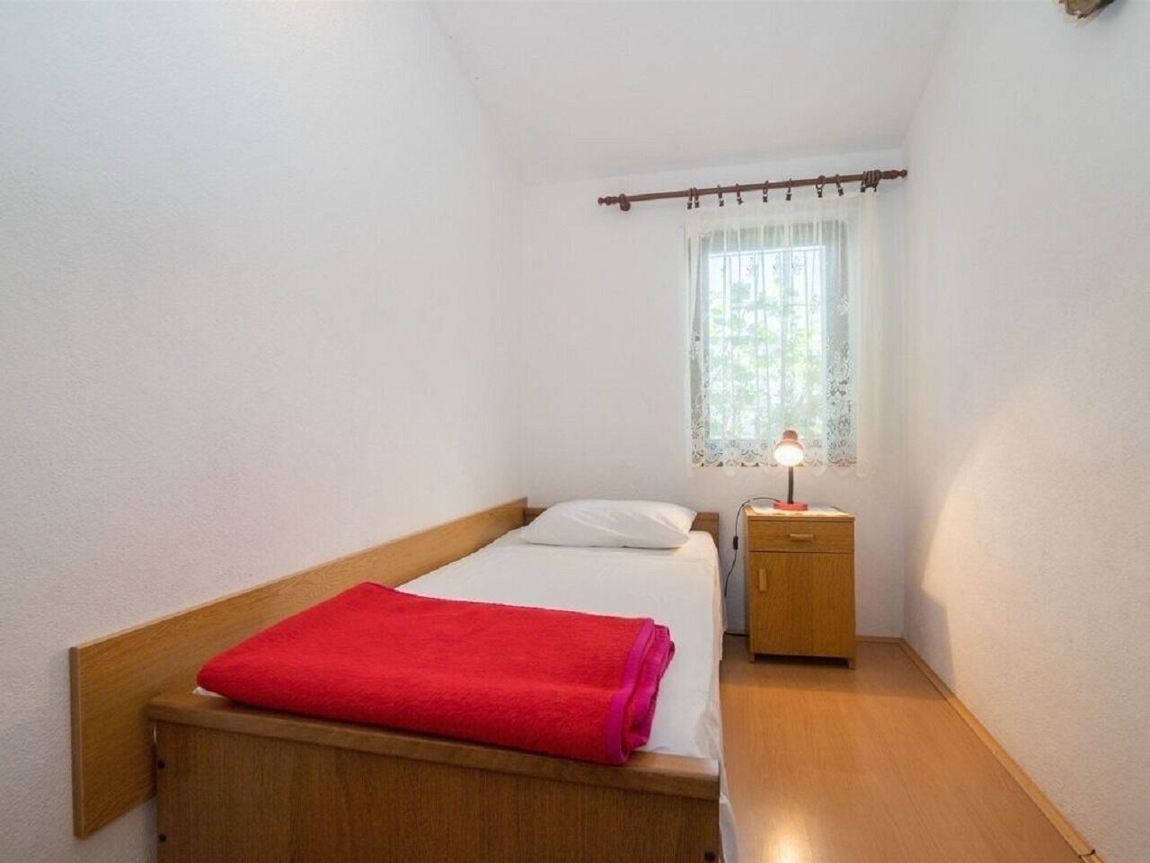 A small, single-bed room with a wooden headboard, a bedside table with a lamp, and a window with a curtain.