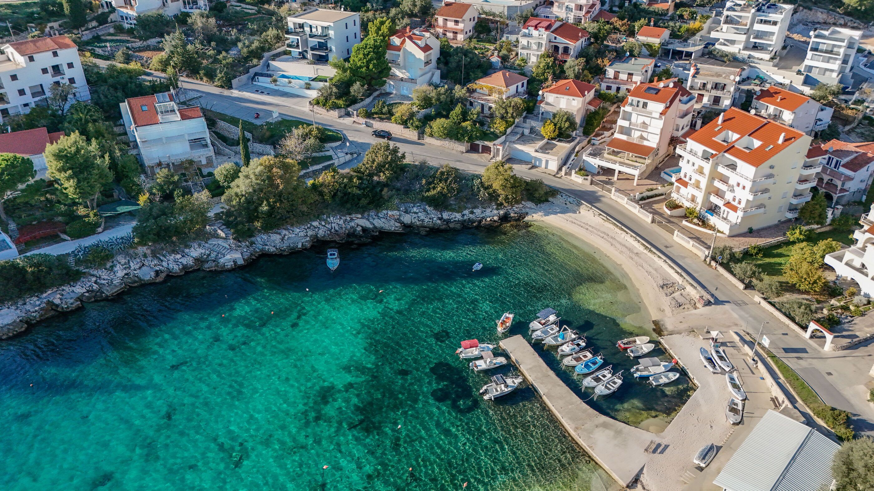 An aerial view of a coastal town with clear turquoise waters, a marina with several boats, and residential buildings with red-tiled roofs.