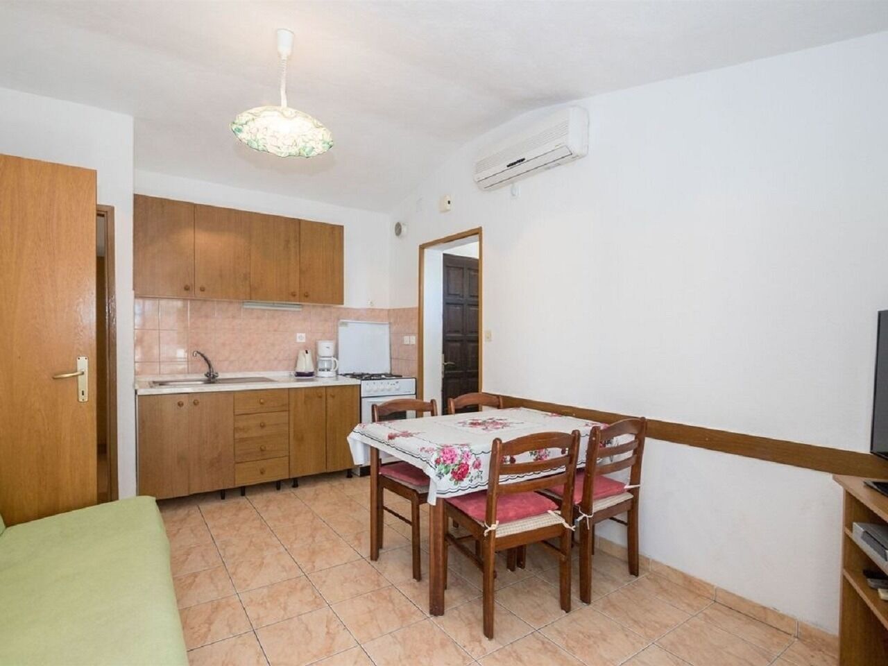 A compact kitchen with wooden cabinets, a dining table with chairs, a small refrigerator, and a ceiling light fixture.