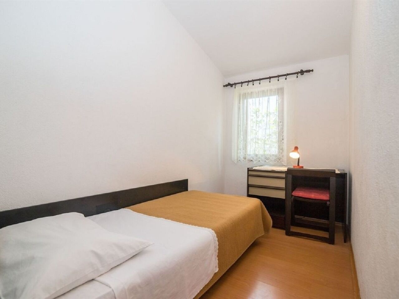 A small, simple room with a single bed, a desk with a chair, and a window with a curtain.