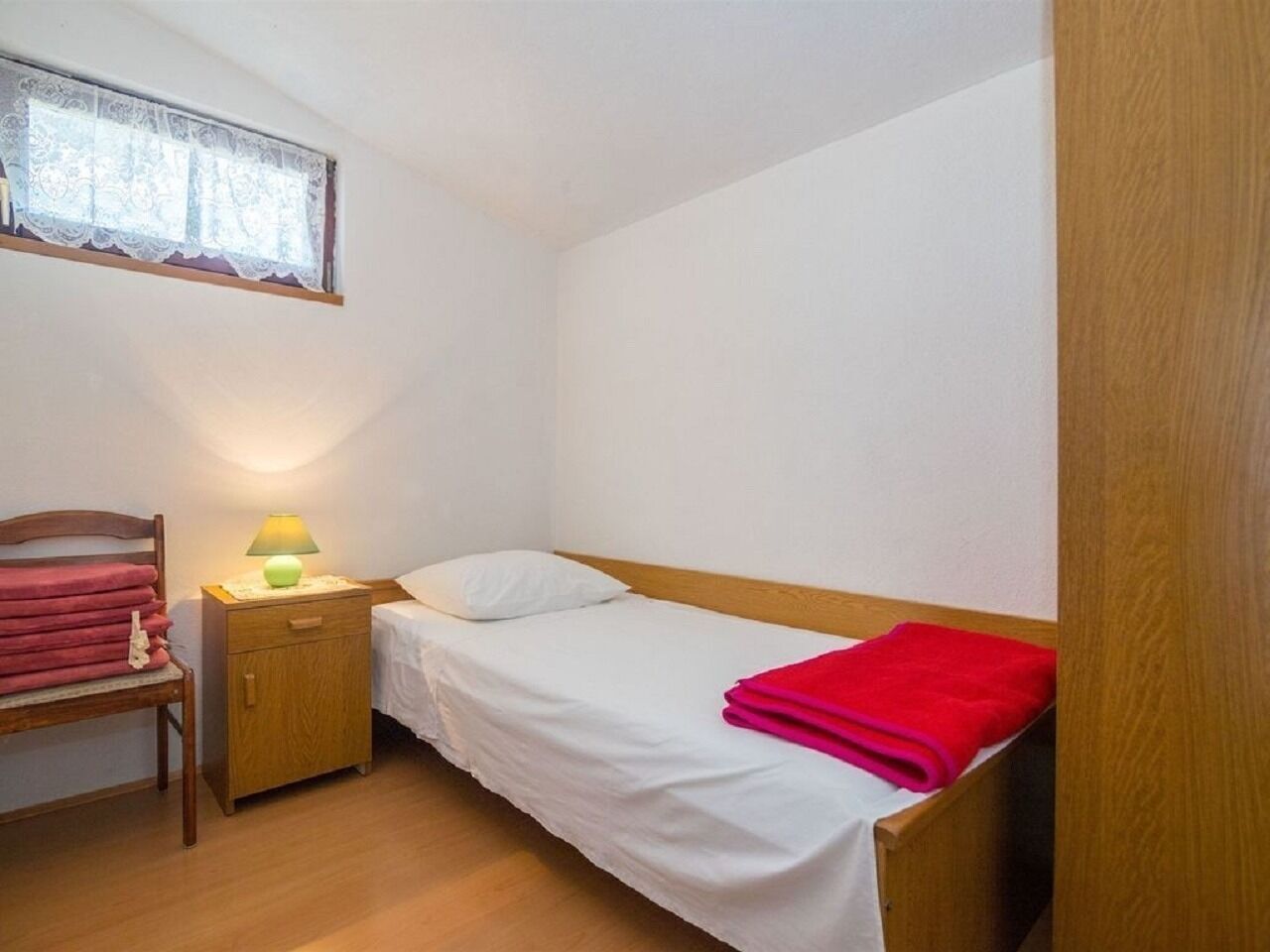 Ein kleines Zimmer mit einem Einzelbett, einem Holzkopfteil, einem Nachttisch mit Lampe, einem Stuhl mit rotem Handtuch und einem Fenster mit Spitzenvorhängen.