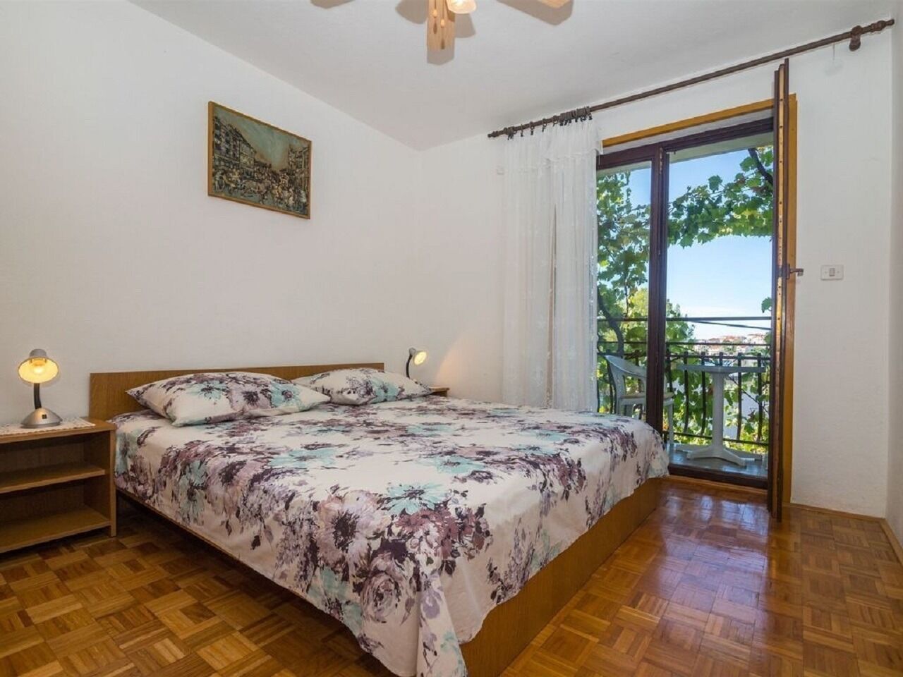 A bedroom with a wooden floor, a bed with floral bedding, a nightstand with a lamp, and a balcony with a view of greenery.