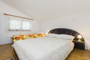A bedroom with a bed, a window with lace curtains, a bedside table with a lamp, and a herringbone-patterned floor. - Apartments Milans (Rogoznica)