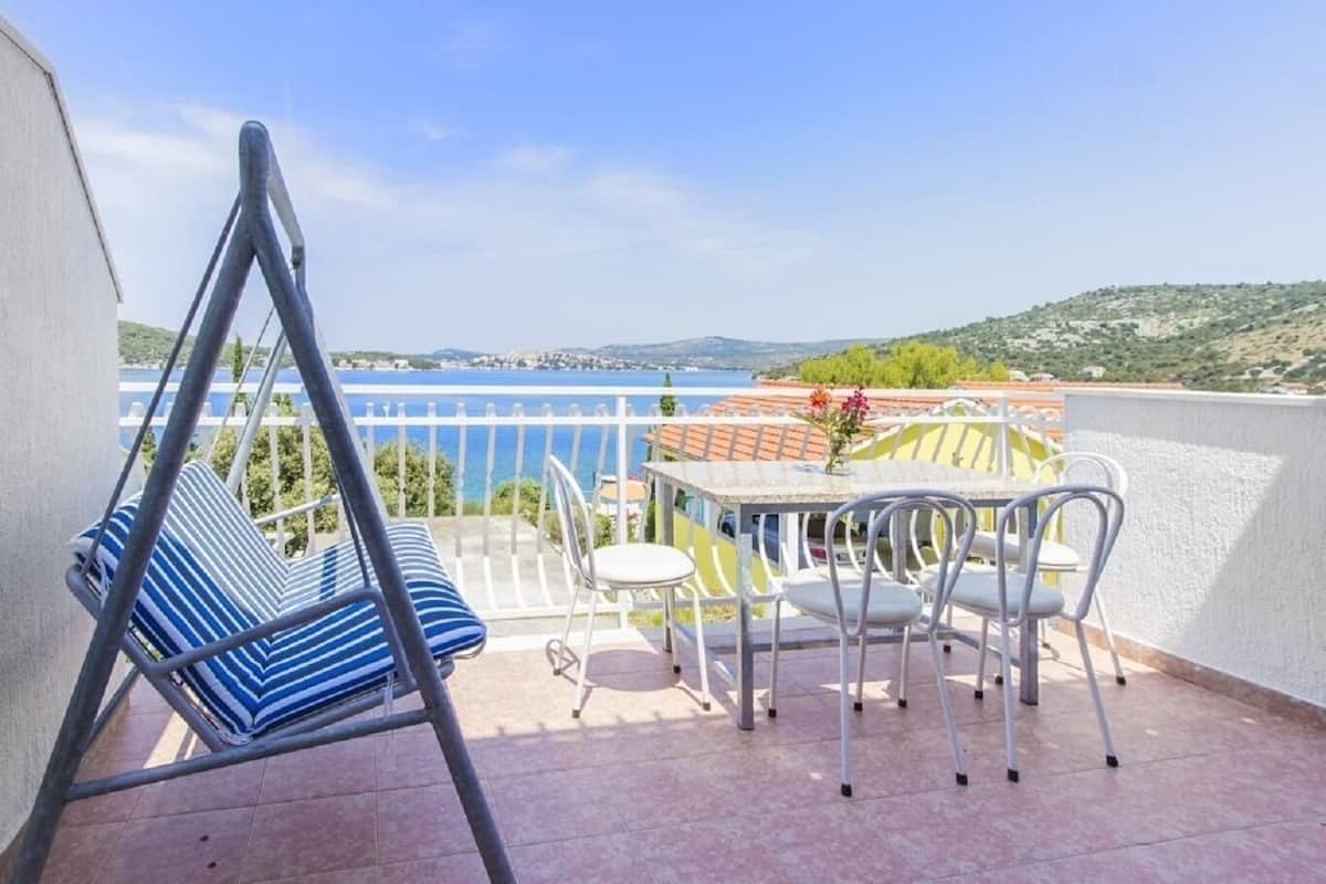 A balcony with a swing chair, a table with chairs, and a view of the sea and hills.