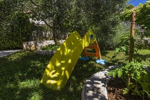A playground with a slide and climbing structure in a grassy area with trees and a stone wall. - Apartments Milans (Rogoznica)