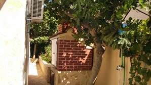 A narrow stone pathway leading to a small brick building with a red door, surrounded by greenery and a metal fence. - Apartment Dusko (Obrovac)