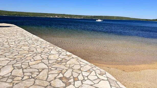 A stone-paved walkway leading to a clear blue sea with a boat in the distance and a green landscape in the background. - Apartment Dusko (Obrovac)
