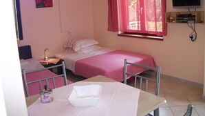 A small, neatly arranged bedroom with a single bed, a bedside table, a desk, and a chair. There is a window with red curtains, a TV mounted on the wall, and a painting on the wall. - Apartment Dusko (Obrovac)