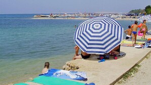 A beach scene with a striped umbrella, people sunbathing, and boats in the background. - Apartments Adriatic (Razanac)