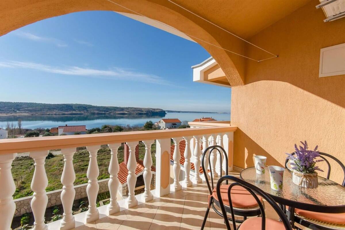 En balkon med udsigt over en sø og fjerne bakker, med hvidt gelænder, et bord med stole og en vase med blomster.