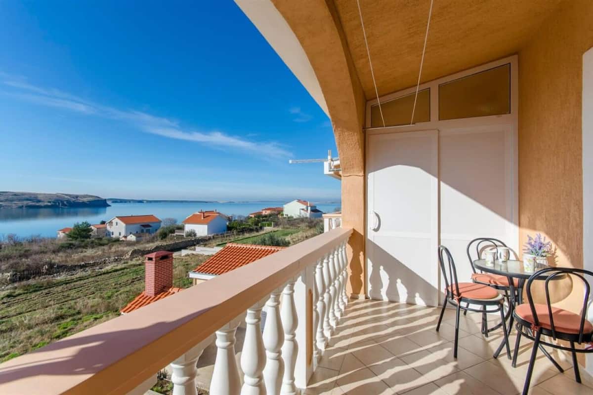 En balkon med udsigt over en sø og huse, et spisebord med stole, og en hvid dør.