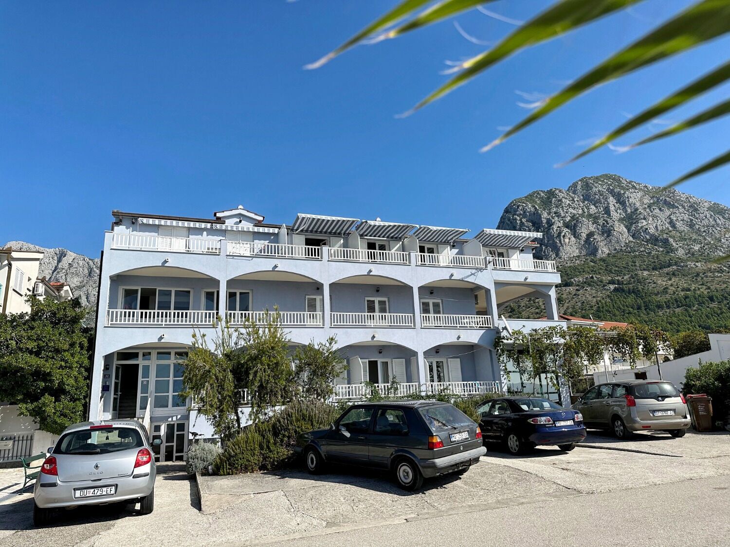 A multi-story building with balconies, parked cars, and a mountainous backdrop.