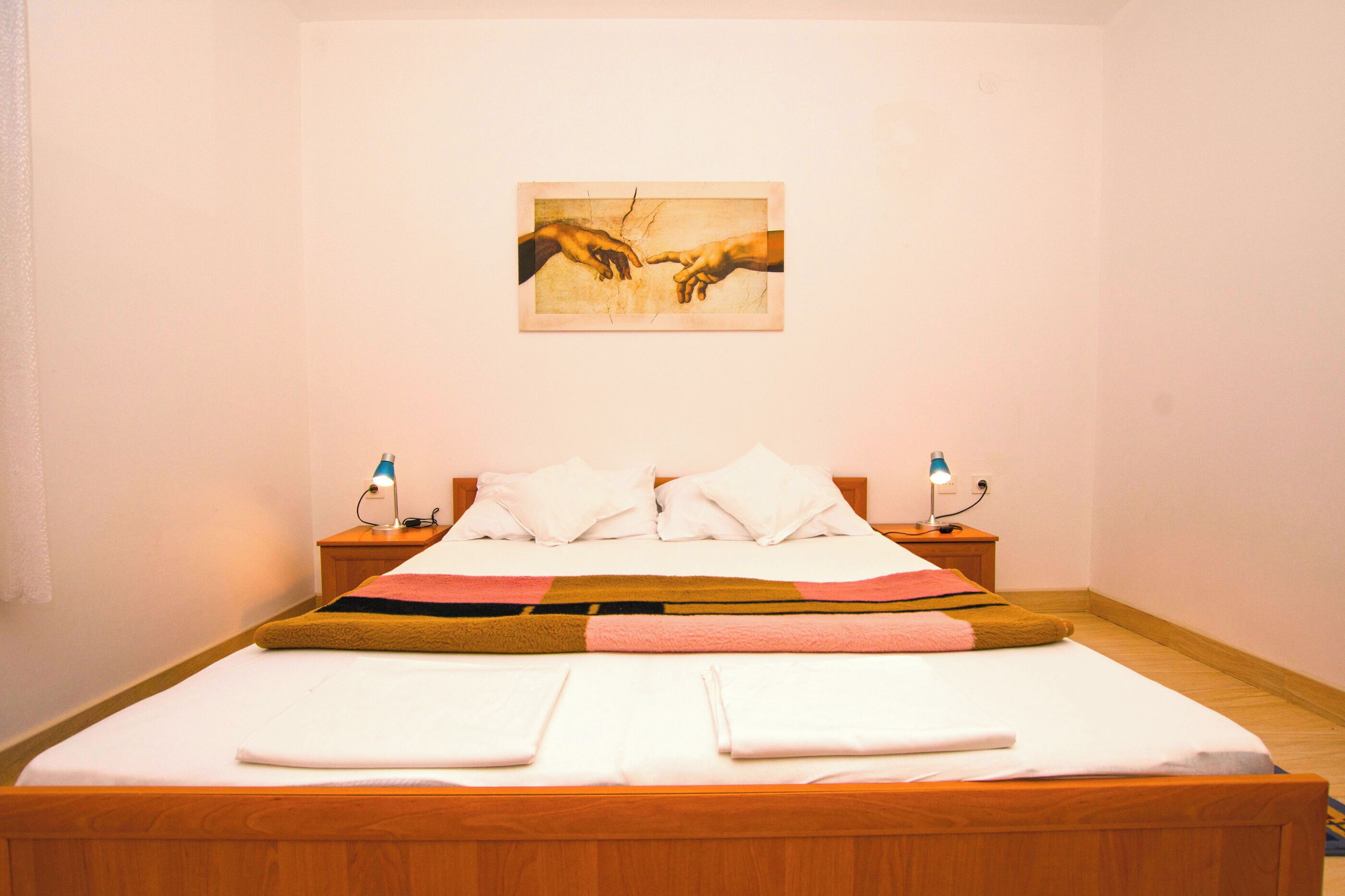 A neatly made bed with white linens and a colorful blanket, two bedside lamps, and a painting of hands on the wall.