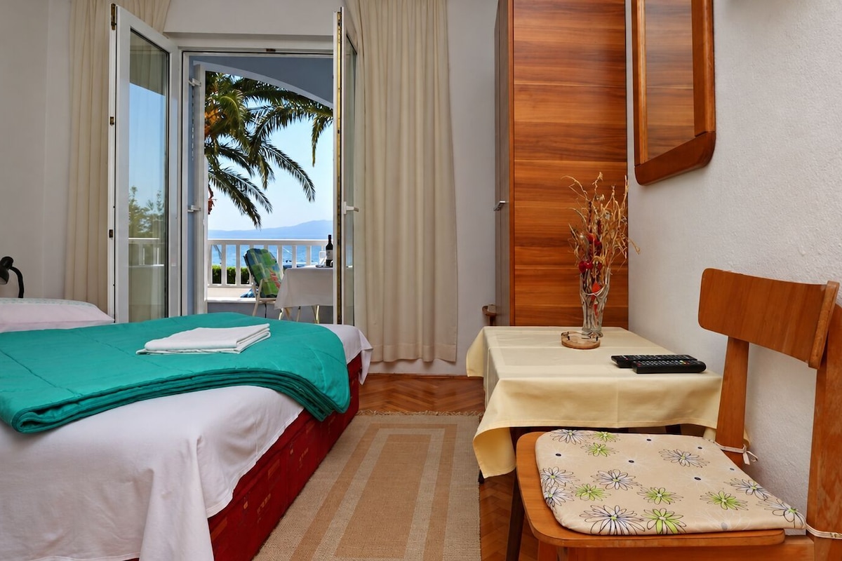 A hotel room with a bed, a small table with a vase, a chair, and a view of the sea through a sliding door.