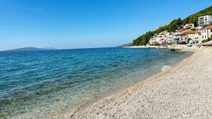 A pebble beach with clear water, buildings on a hillside, and mountains in the distance. - Apartments Tomo (Gradac)
