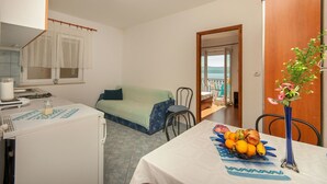 Una habitación compacta con kitchenette, un comedor con frutero, un sofá verde y vista al mar a través de una puerta abierta.