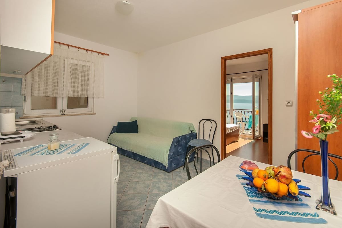 Une pièce compacte avec une kitchenette, un coin repas agrémenté d’un panier de fruits, un canapé vert et une vue sur la mer par une porte ouverte.