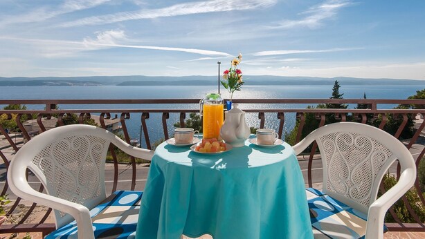 Una mesa con una jarra de jugo de naranja, un florero con flores y un plato de fruta en un balcón con vistas al agua y colinas distantes.