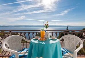 Una mesa con una jarra de jugo de naranja, un florero con flores y un plato de fruta en un balcón con vistas al agua y colinas distantes.