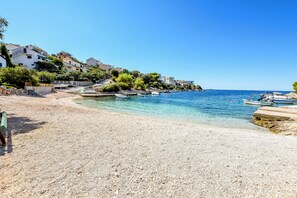 Egy kavicsos strand, ahol csónakok horgonyoznak, a víz kristálytiszta, és az épületek egy lejtős domboldalon állnak.