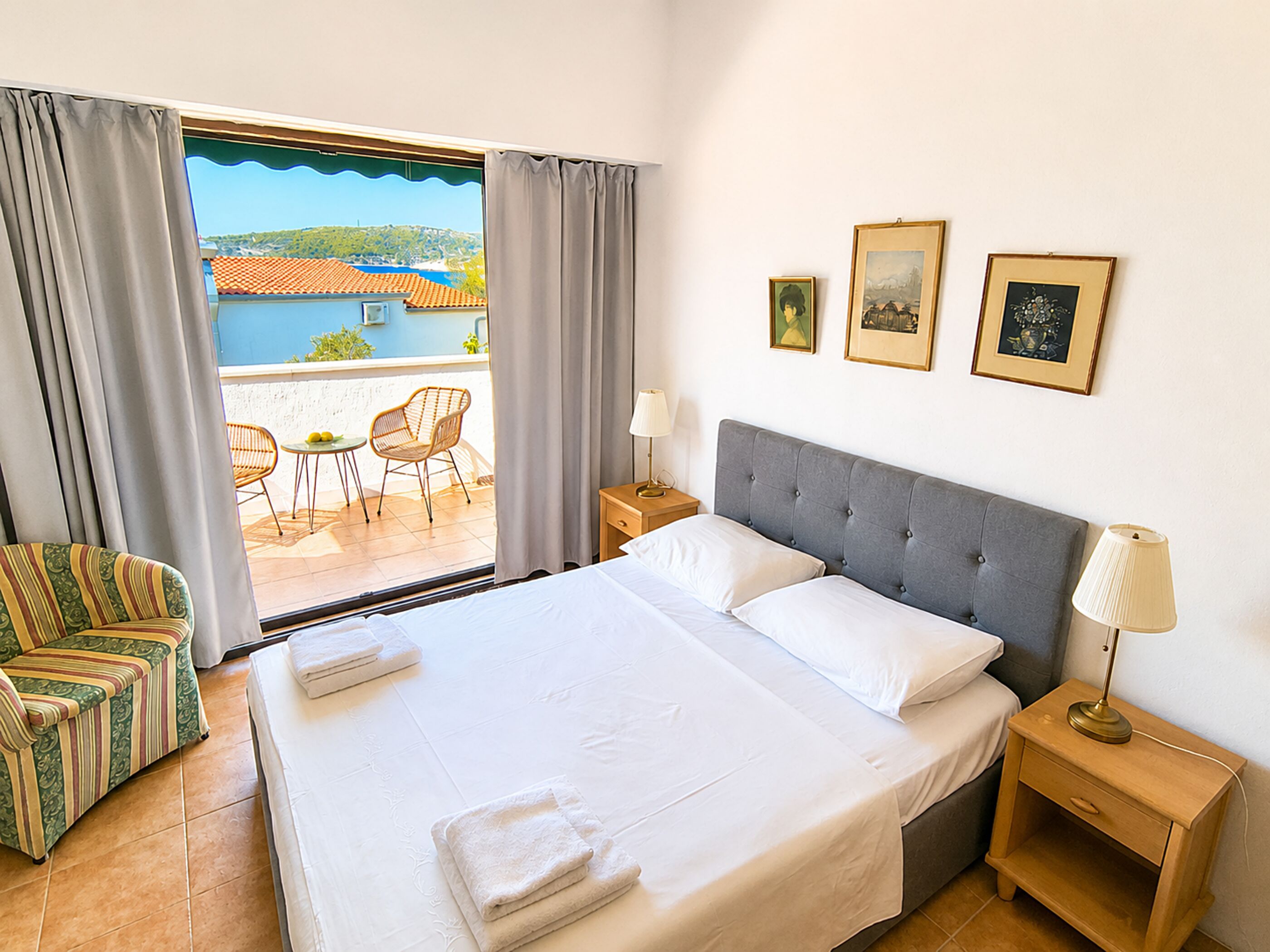 A hotel room with a bed, a nightstand, a chair, a table, and a view of the sea through large windows.
