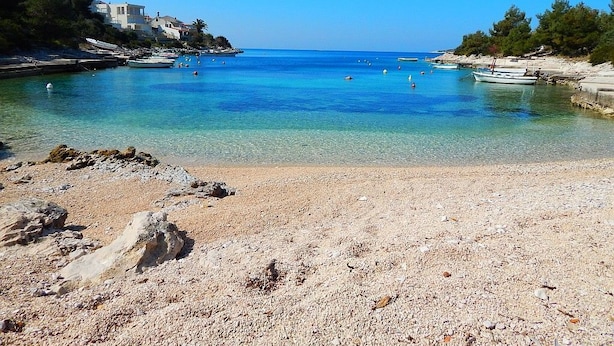 Una spiaggia di ciottoli con acque cristalline, imbarcazioni e edifici sullo sfondo.