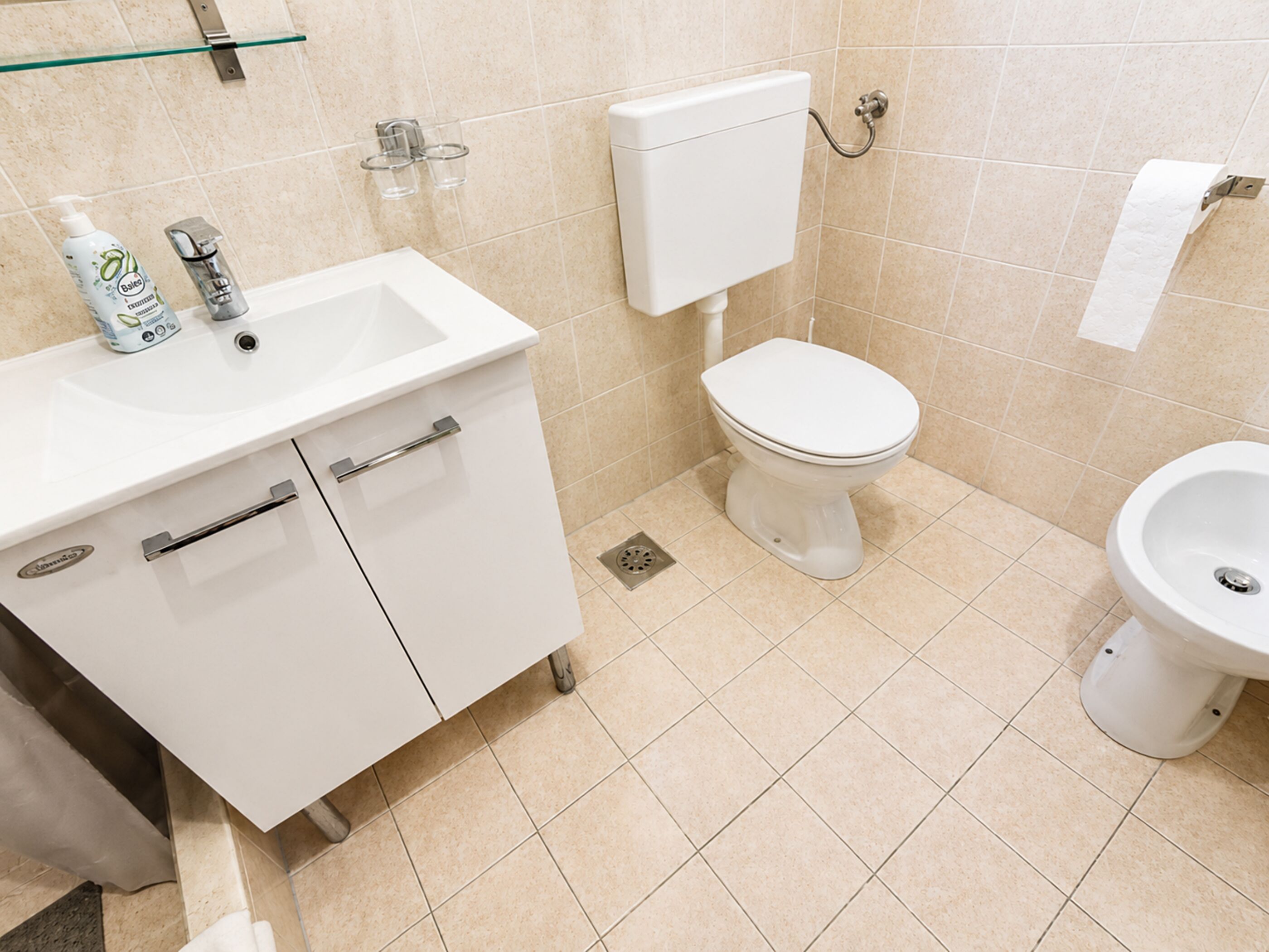 A bathroom with a white sink, toilet, and bidet. There is a glass shelf above the sink and a roll of toilet paper on the wall.