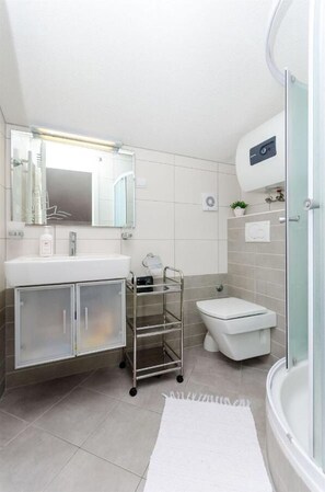 Een moderne badkamer met een witte wastafel, spiegel en toilet. Er is een douchecabine met glazen wanden en een witte vloerkleed.