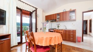 A dining area with a table and chairs, a kitchen with wooden cabinets, and a view through a glass door. - Apartment Ivan (Sali)