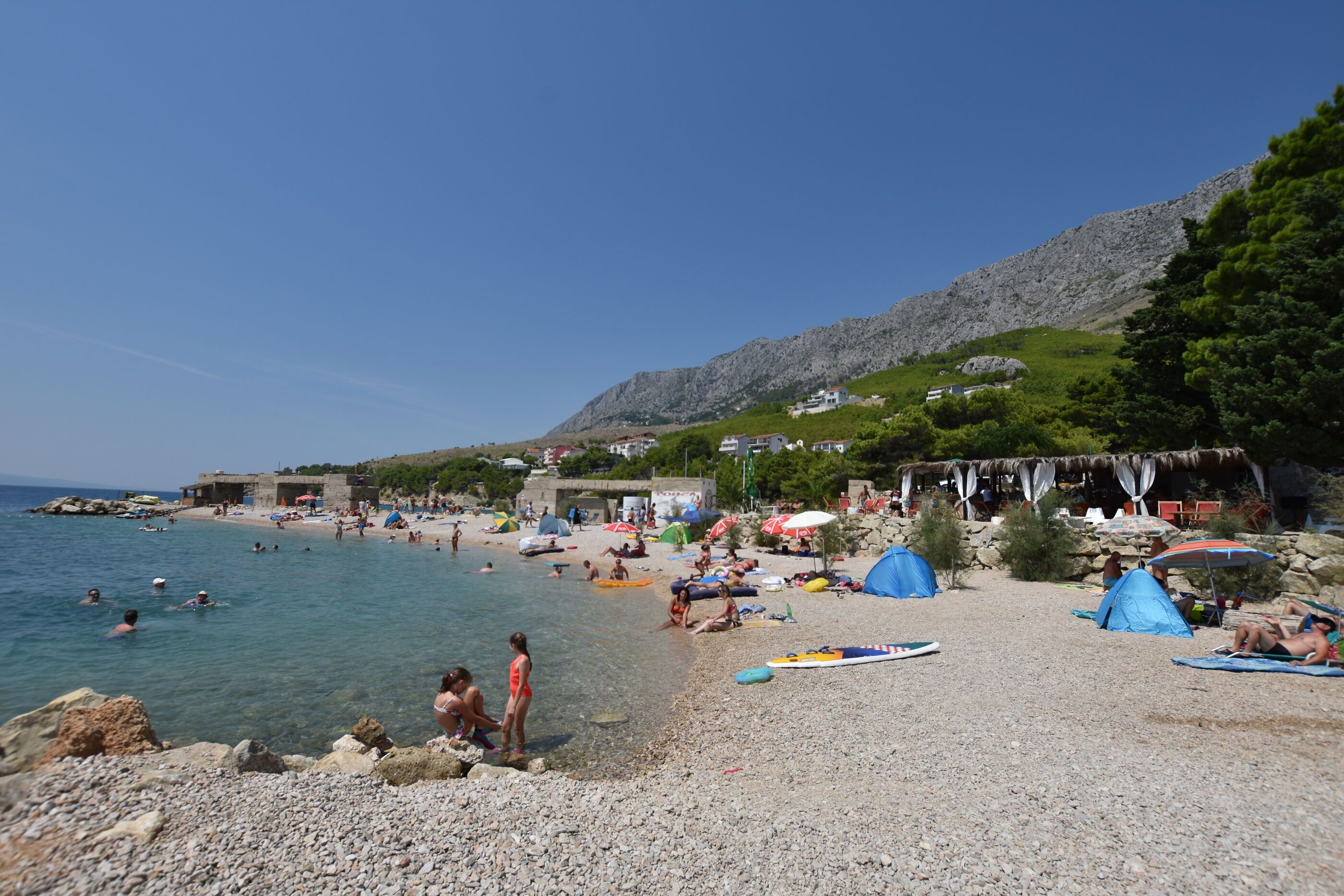 A pebble beach with clear water, tents, and people.