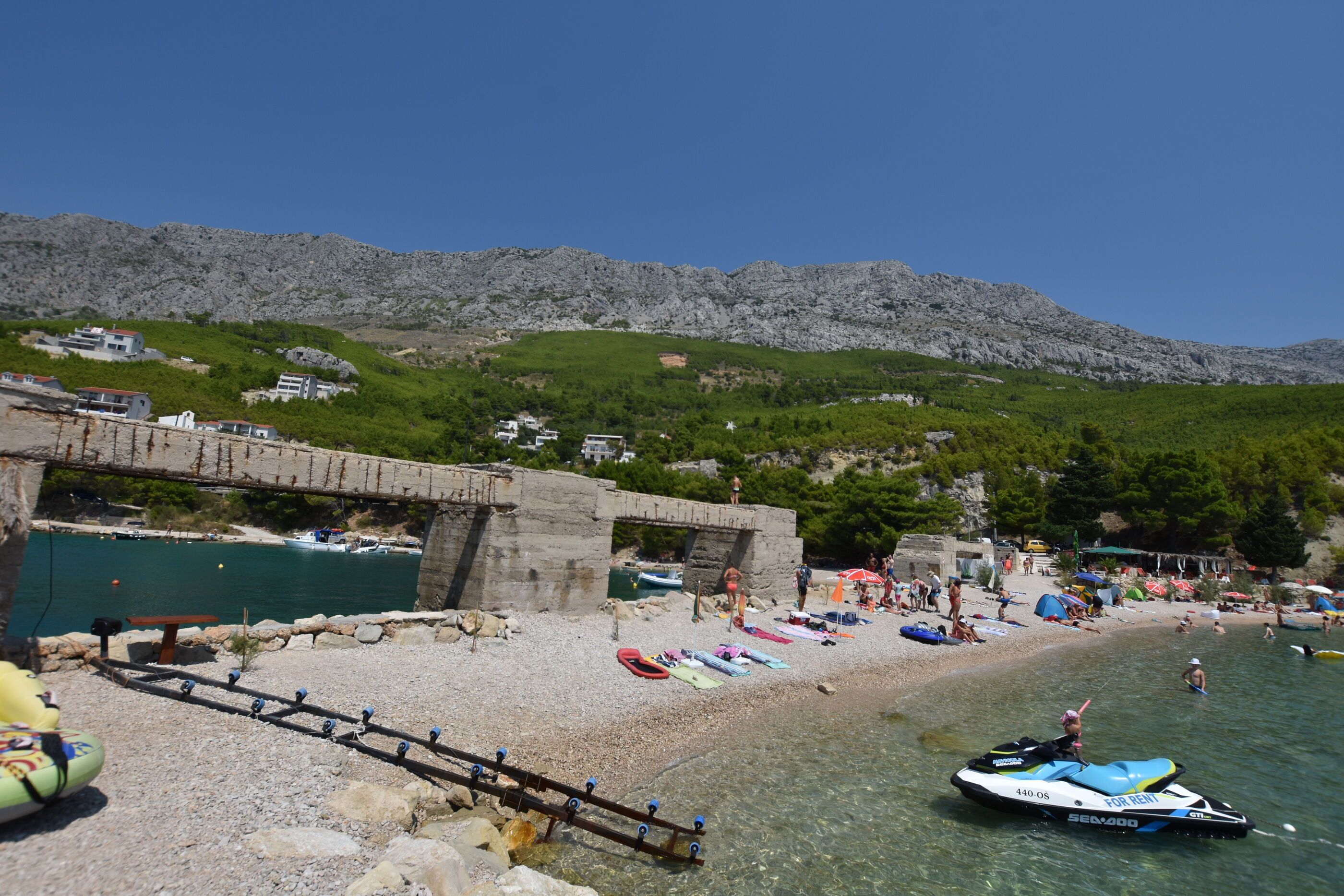 A beach with boats, a jet ski, and a stone bridge.