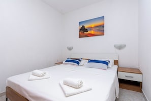 A neatly made bed with white linens and two towels, a bedside table with a lamp, and a painting of a sunset on the wall. - Apartments Marko (Omis)